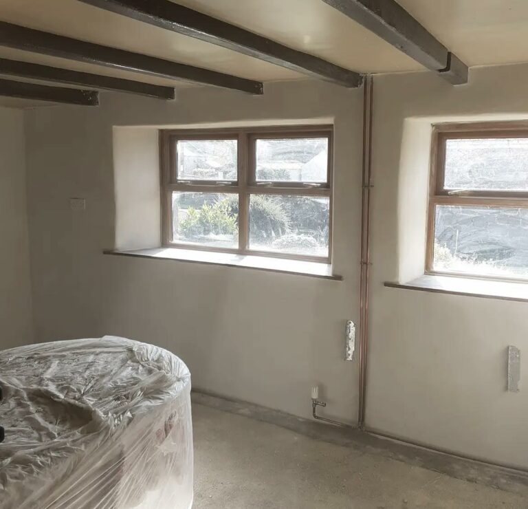 A room with a covered piece of furniture, possibly during renovation. Natural light streams through two small-paned windows with a white sill, contrasting with the dark ceiling beams above. The walls have been plastered with lime plastering.